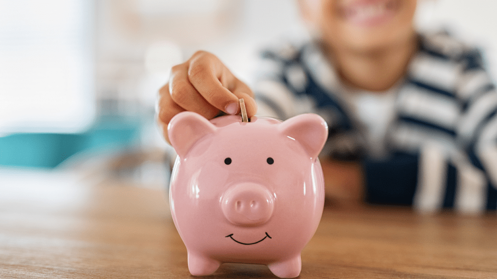 menino colocando uma moeda em um cofrinho com o formato de um porquinho cor de rosa