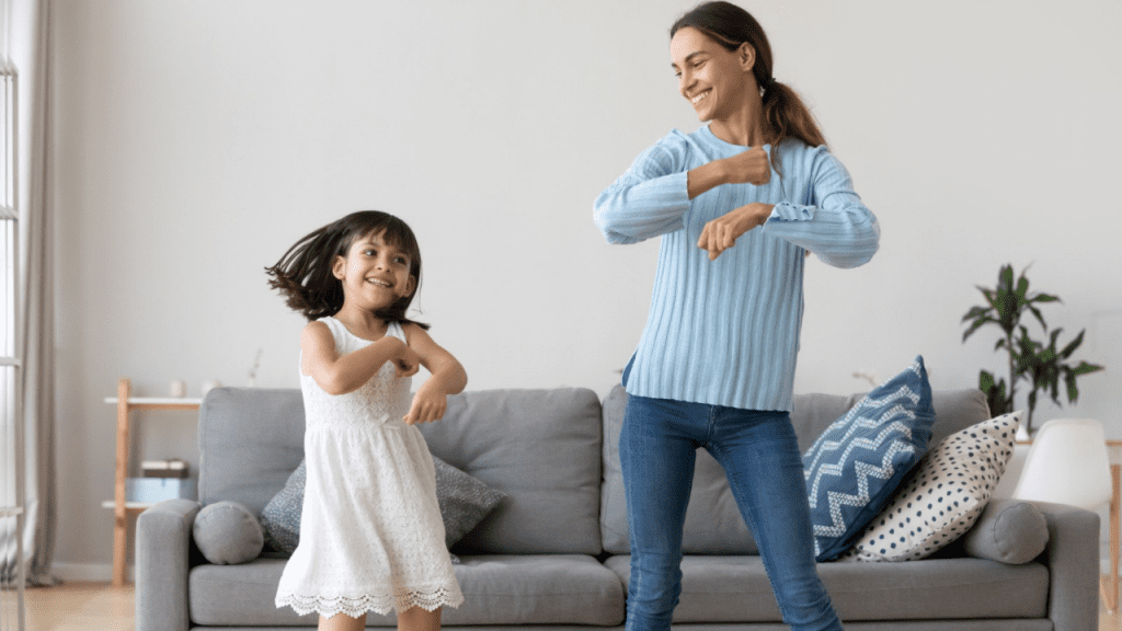 mulher vestindo calça jeans e blusa azul está dançando ao lado de uma menina de vestido branco. As duas estão na frente de um sofá cinza, sob o qual tem duas almofadas azuis com listras brancas. Ao fundo, uma parede branca e uma planta.