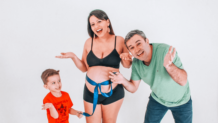 Imagem mostra uma mulher grávida esperando o segundo filho com um laço azul na barriga. De um lado dela, está o primeiro filho e do outro ládo está o marido. Os dois com as mãos na barriga dela.