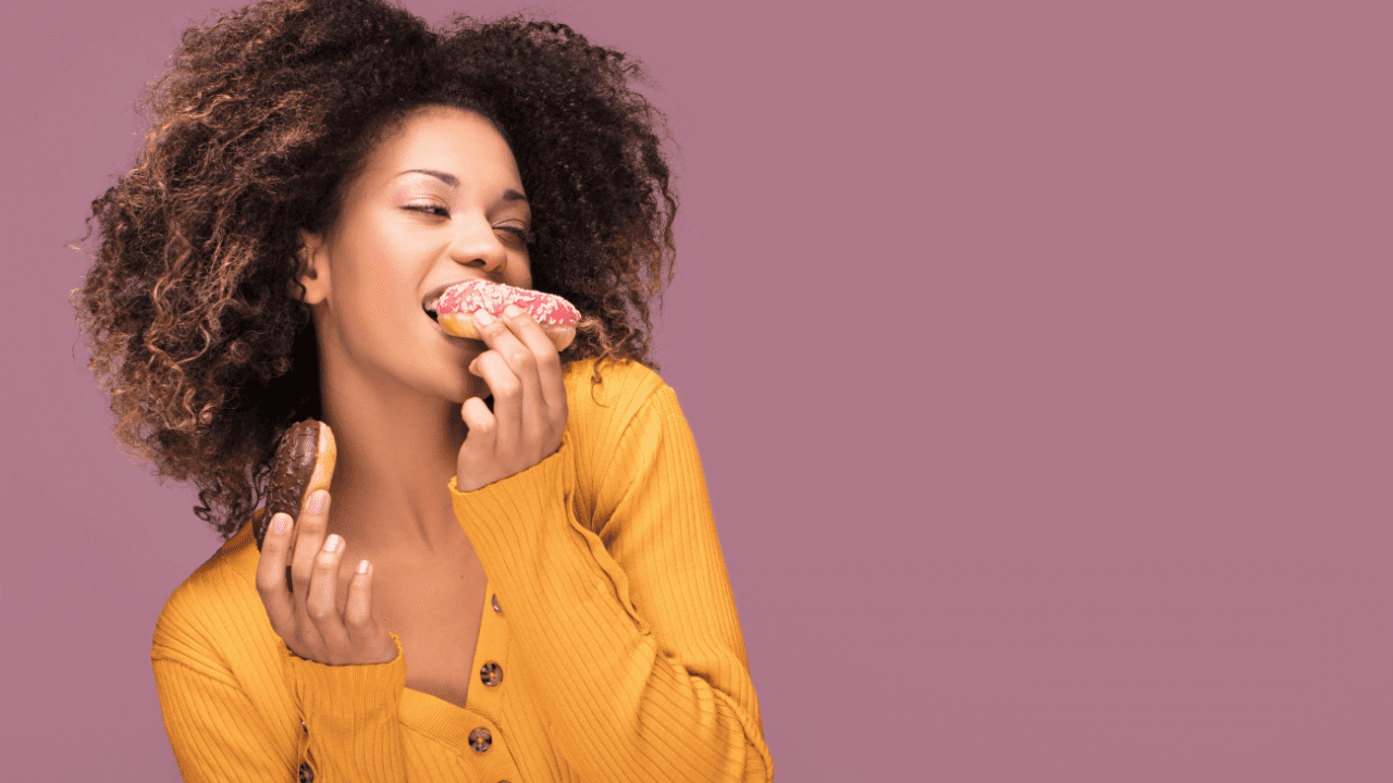 uma mulher negra com camisa amarela e cabelos crespos e deourados está comendo um donnut, para ilustrar o conteúdo sobre comer emocional